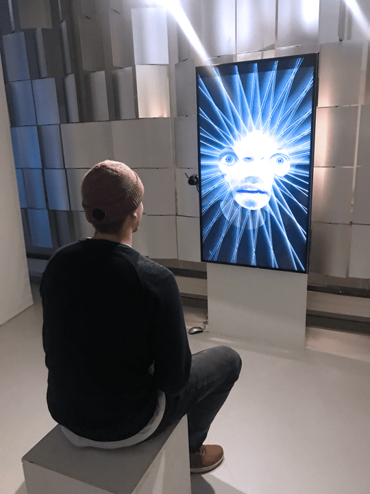 A visitor sits in front of the "Expression Mirror" that presents the detected emotions as a collage of different faces.